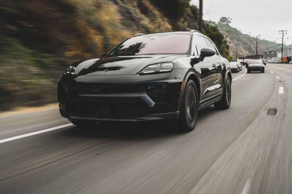 A porsche macan driving along a mountainside with electricity poles in the background. There are other Porsche cars following behind