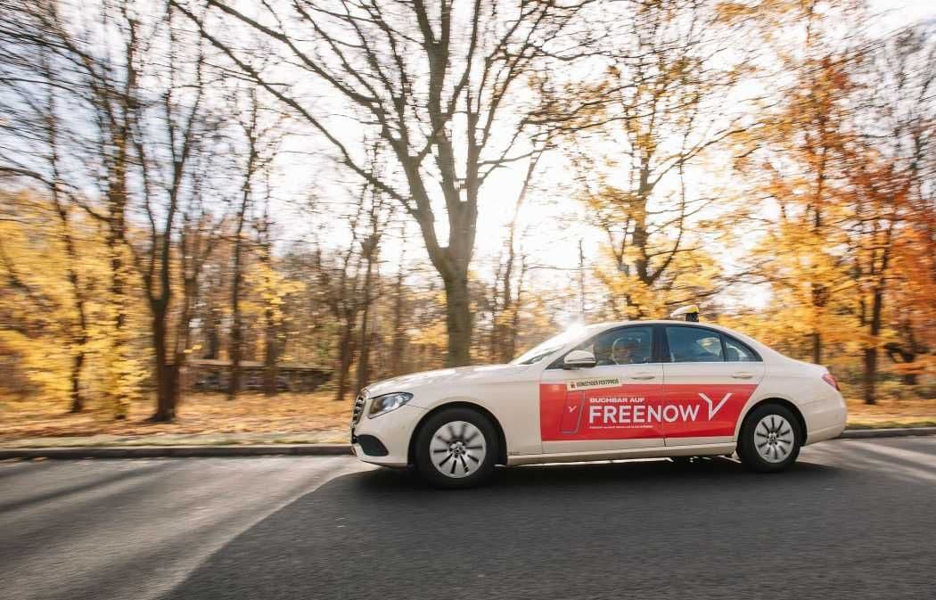 Weißes Auto mit dem Namen des Taxiunternehmens „Free Now“ an der Seite, das unter einem klaren Himmel auf einer von Herbstbäumen gesäumten Straße fährt.