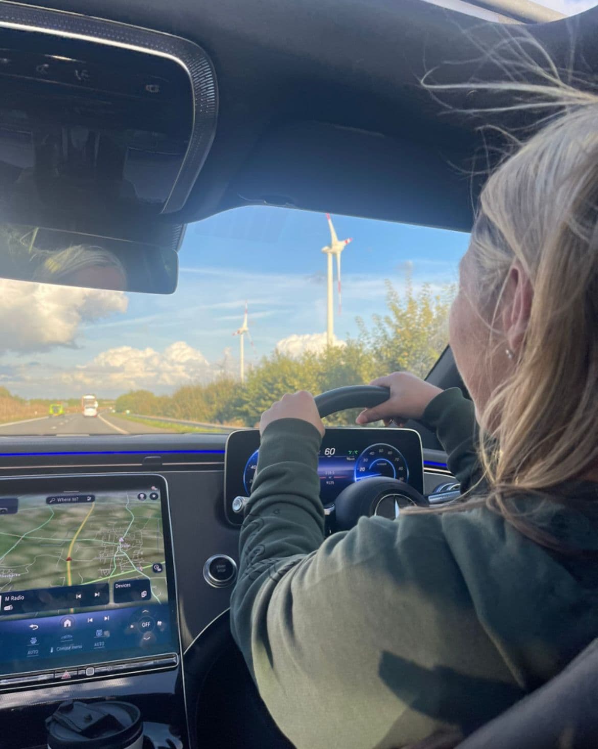 Amanda Armitage driving an EV with wind turbines outside. Amanda Armitage conduisant un véhicule électrique avec des éoliennes à l'extérieur. Amanda Armitage fährt ein Elektrofahrzeug mit Windturbinen im Außenbereich. Amanda Armitage a conduzir um veículo elétrico com turbinas eólicas no exterior. Amanda Armitage alla guida di un veicolo elettrico con turbine eoliche all'esterno. Amanda Armitage conduciendo un vehículo eléctrico con turbinas eólicas en el exterior.