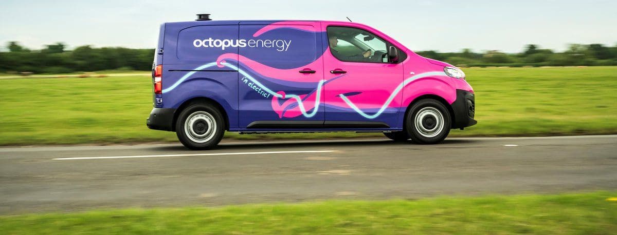Furgoneta Octopus Energy de color rosa y morado circulando por una carretera rural con campos verdes al fondo.
