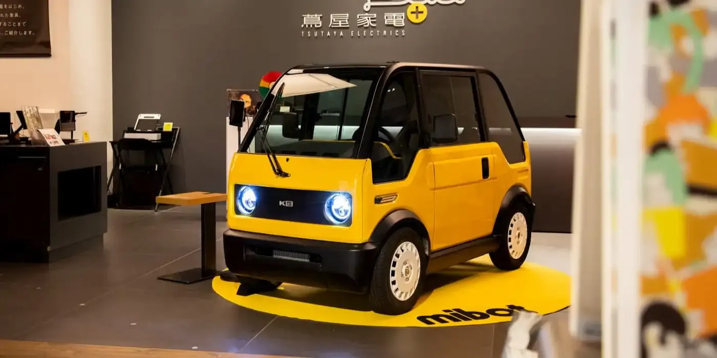 Ein kompaktes gelbes Elektroauto mit runden Scheinwerfern, das in einem Innenbereich auf einer runden Plattform ausgestellt ist und von modernem Dekor umgeben ist.