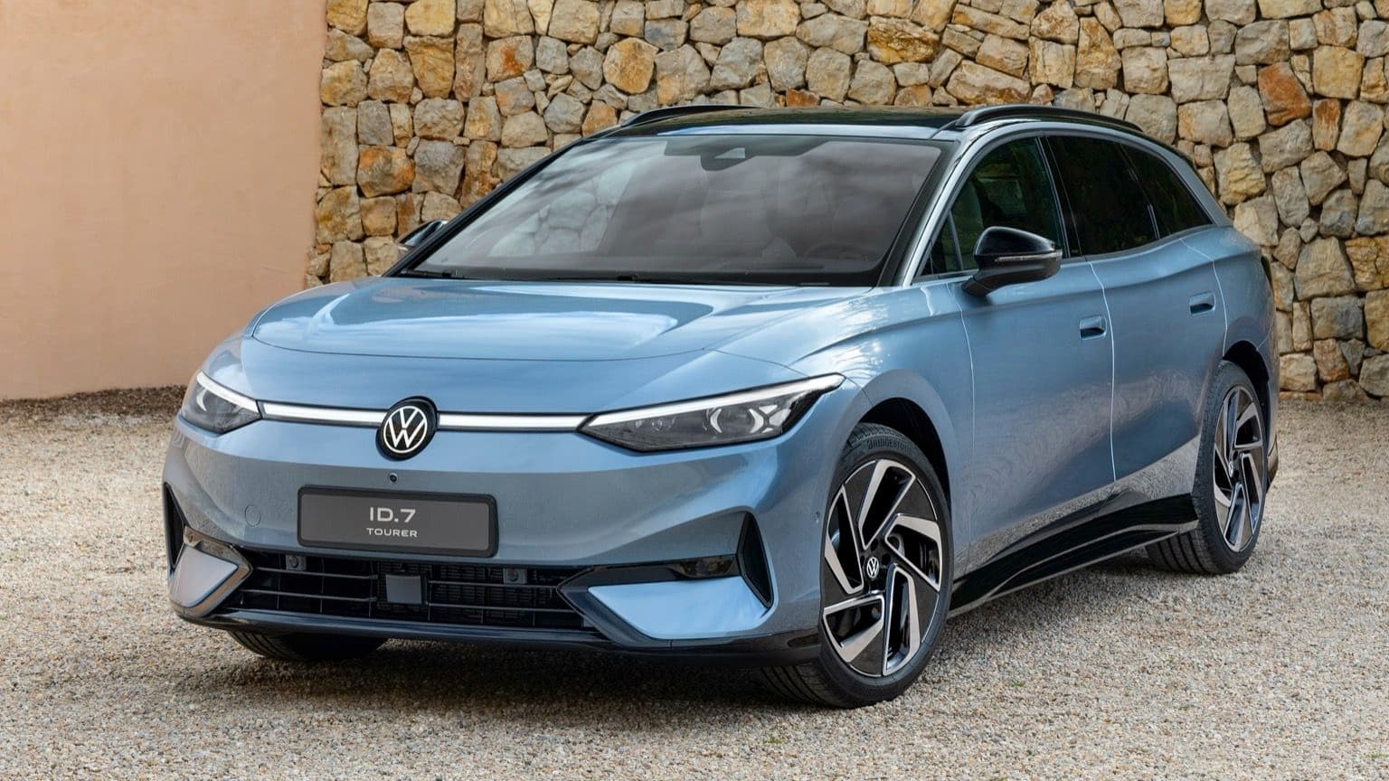 Volkswagen ID.7 Tourer azul estacionado sobre grava, con un muro de piedra de fondo. Presenta un diseño elegante y faros modernos.