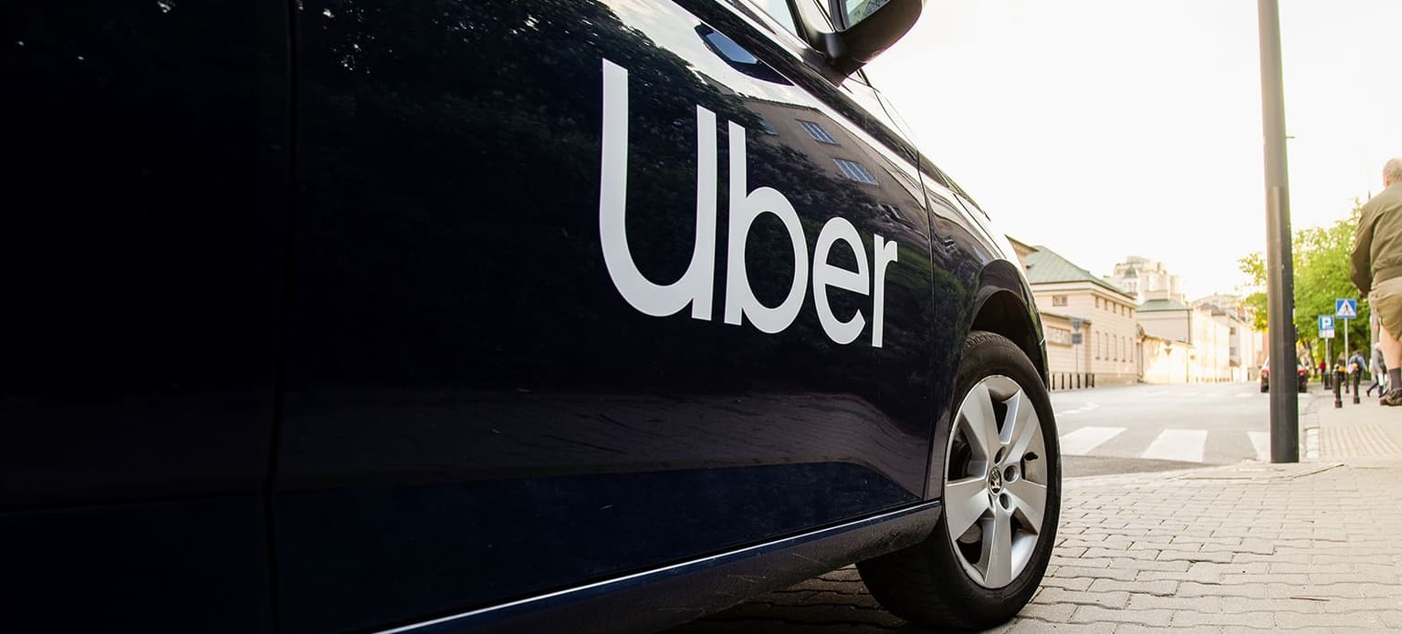 Ein geparktes schwarzes Auto mit dem Uber-Logo an der Seite, auf einer Stadtstraße mit Gebäuden und Bäumen im Hintergrund.