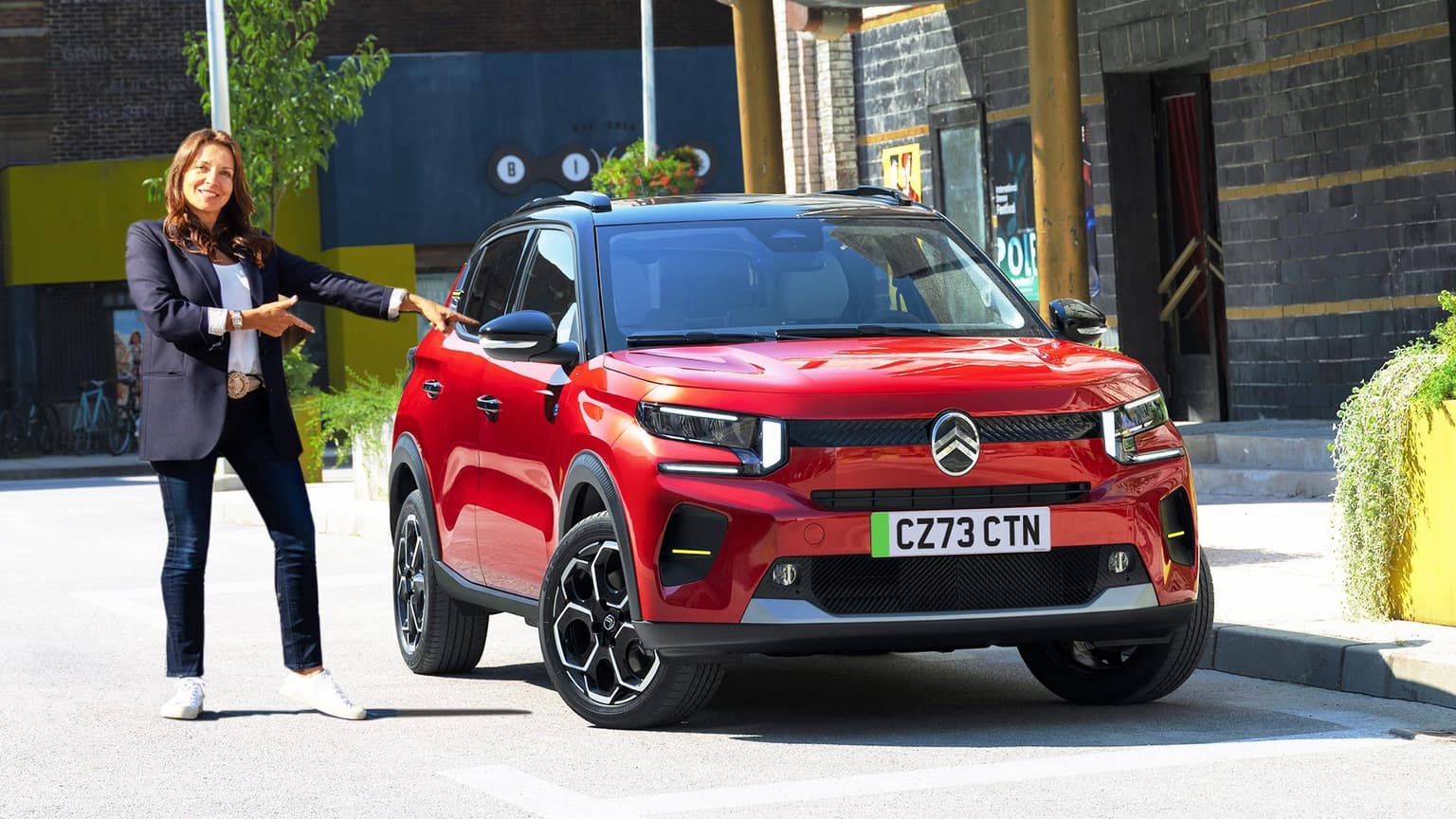 A red Citroen ec3 car parked on a sunny road with a smartly dressed brunette woman pointing at the electric vehicle and smiling