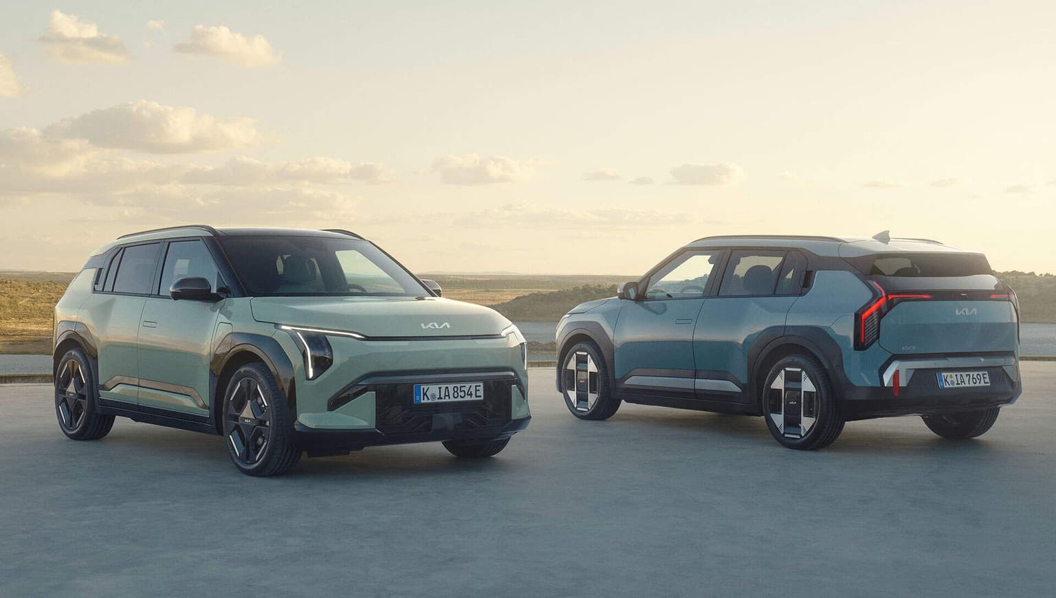 Two KIA EV-3 SUVs in a parking area, one facing forward and the other backward, under a clear sky with a scenic landscape in the background. Due SUV KIA EV-3 in un parcheggio, uno rivolto in avanti e l'altro all'indietro, sotto un cielo limpido con un paesaggio panoramico sullo sfondo.