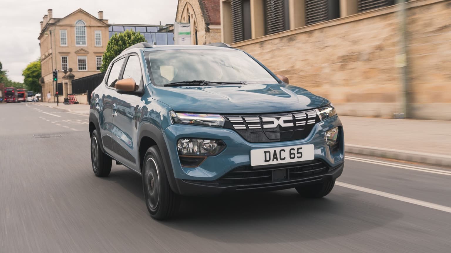 A new dark blue Dacia Spring electric vehicle in motion driving down a quiet road alongside a yellow brick building