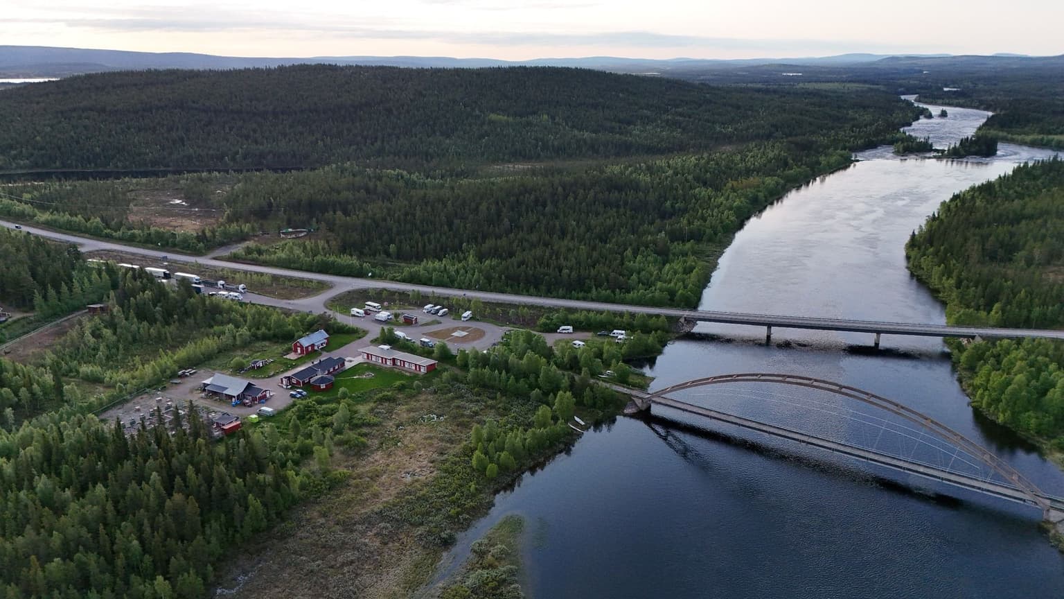 Aerial view of a lush, forested landscape with a river, a bridge, and a small cluster of buildings near a road.