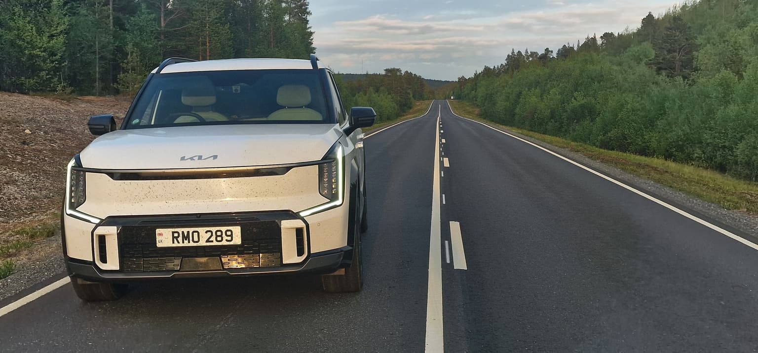 White Kia SUV parked on a deserted, straight road surrounded by lush green forests under a cloudy sky.