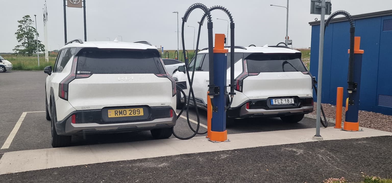 Two white electric Kia SUVs charging at a station, with charging cables connected. Overcast sky and a blue building in the background.