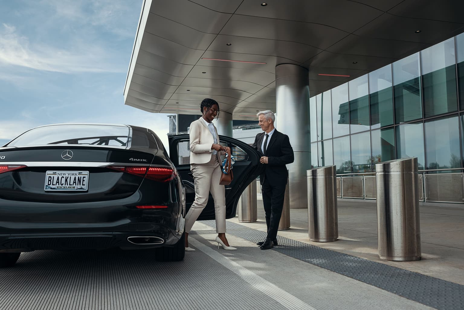 Eine Frau im Kostüm steigt aus einem Premium-Fahrzeug mit dem Kennzeichen „Blacklane“ aus, während ihr ein Mann im schwarzen Anzug die Tür aufhält. Vor einem futuristischen Gebäude, das wie ein Flughafen aussieht.