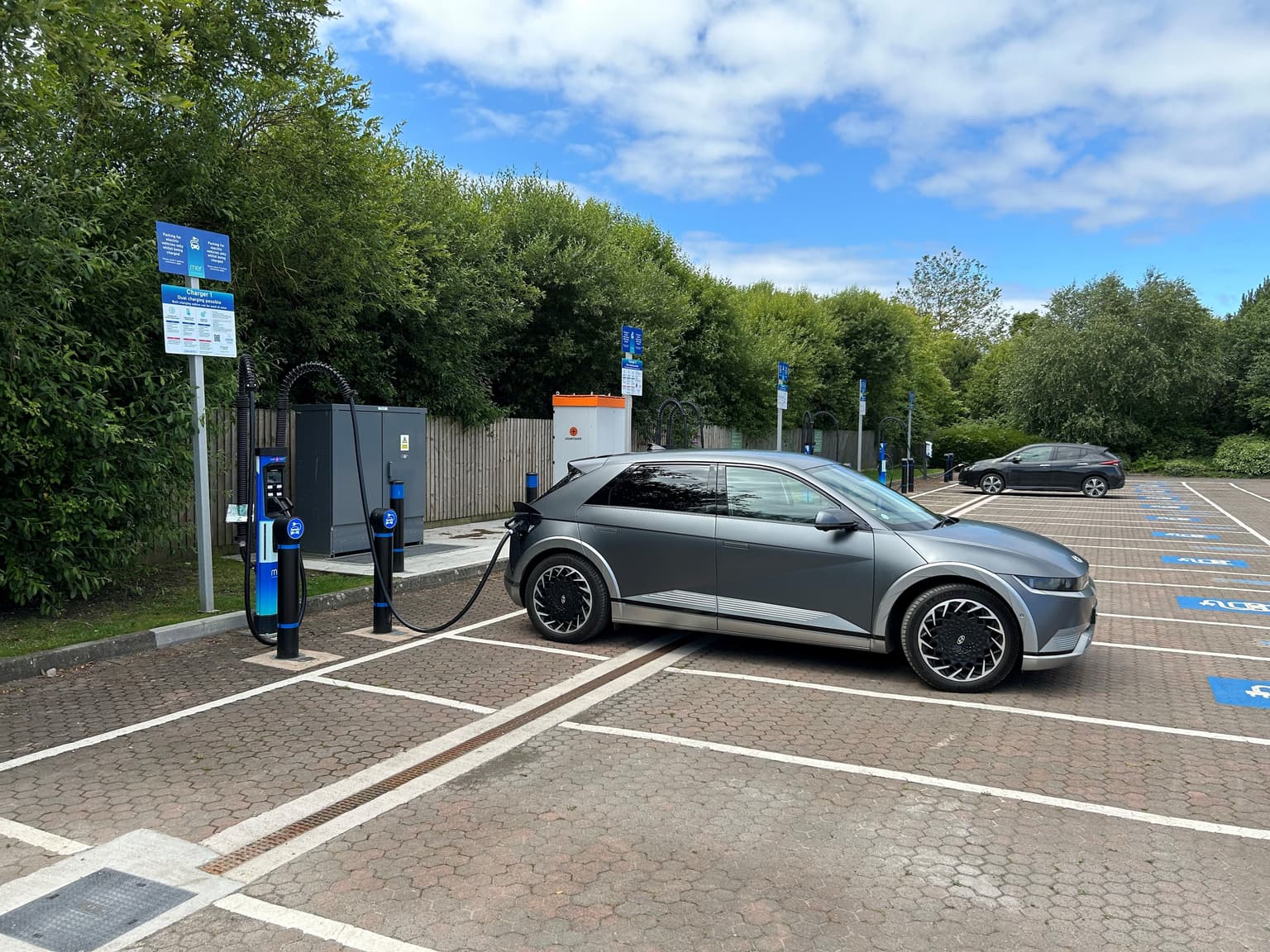 An electric vehicle plugged into a small, blue, Mer charger in a car park with green trees in the background.