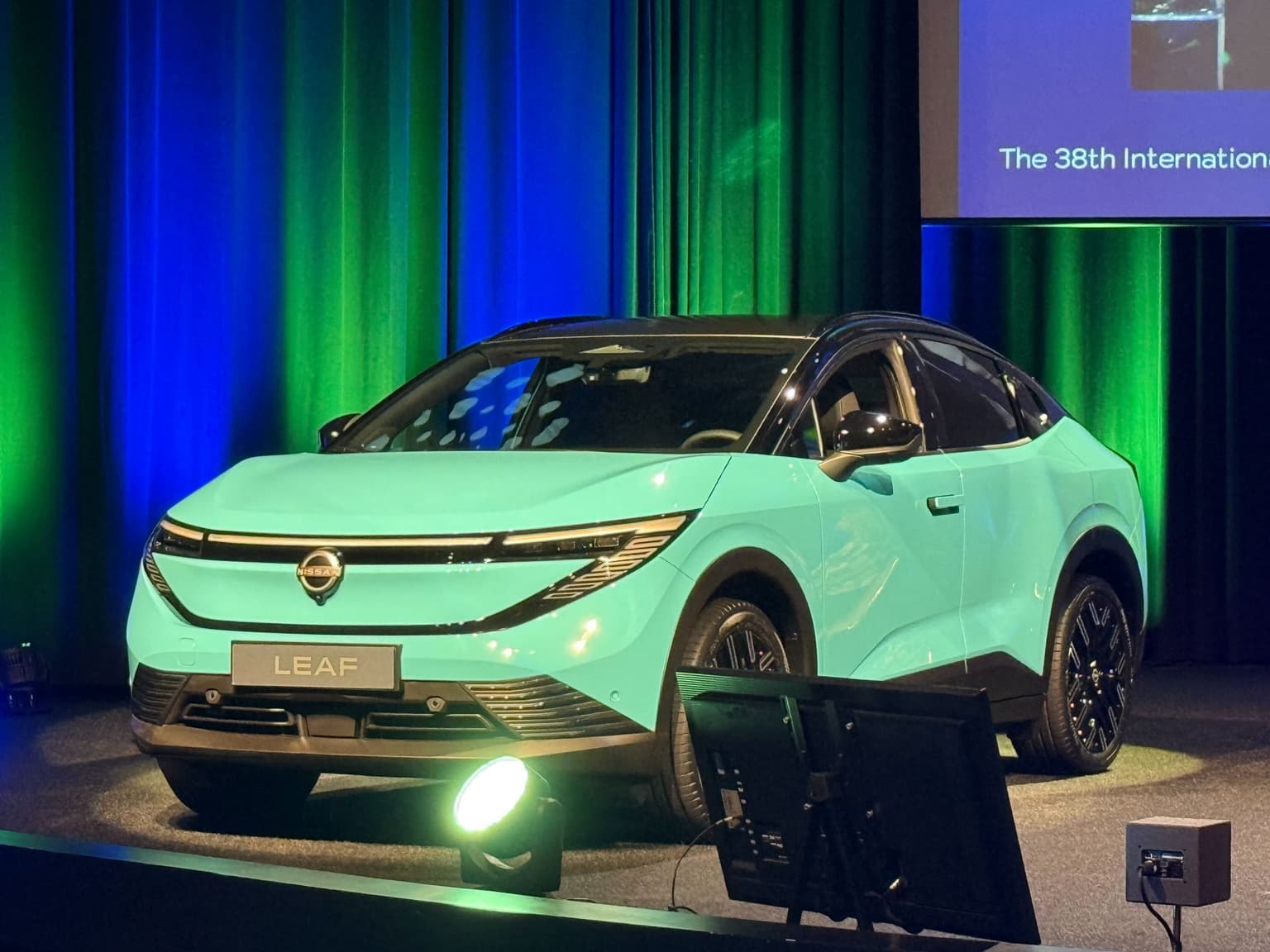 A light green coloured Nissan Leaf 2025 model, with a blue and green background