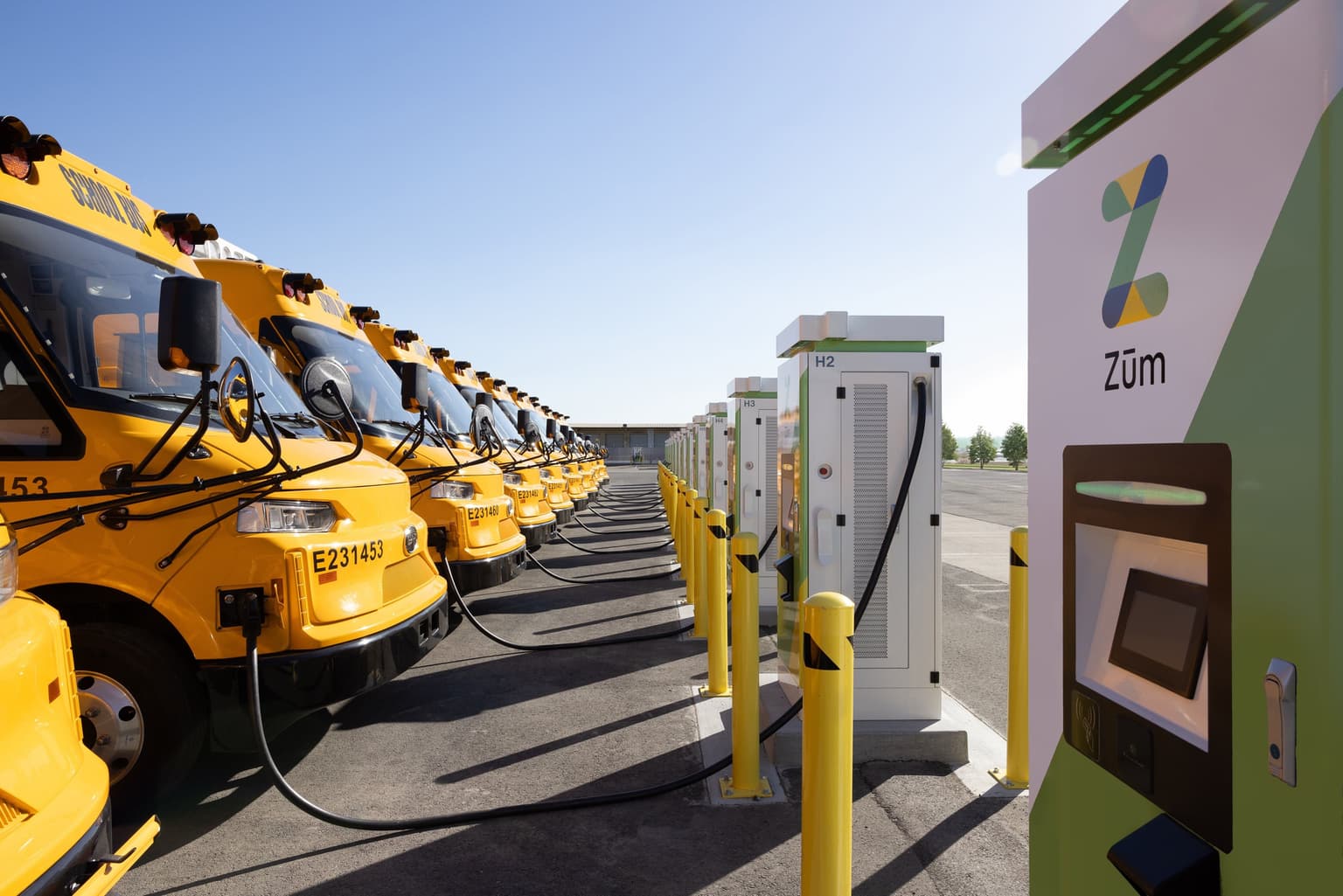 Autobuses escolares amarillos cargándose en estaciones eléctricas en un aparcamiento bajo un cielo azul claro.