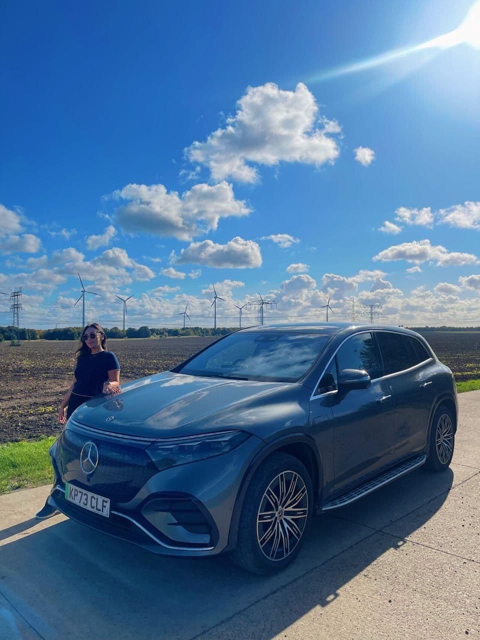 Angela standing next to EQS EV in front of wind turbines. Angela de pie junto a EQS EV frente a las turbinas eólicas. Angela in piedi accanto alla EQS EV davanti alle turbine eoliche. Ângela de pé junto à EQS EV em frente às turbinas eólicas. Angela steht neben dem EQS EV vor Windrädern. Angela debout à côté d'EQS EV devant des éoliennes.