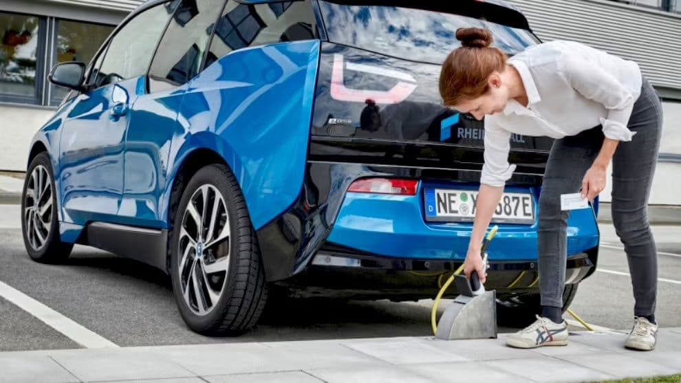 Mujer cargando un coche eléctrico azul en una estación de carga. Sostiene un enchufe de carga, con el coche aparcado en un espacio designado.