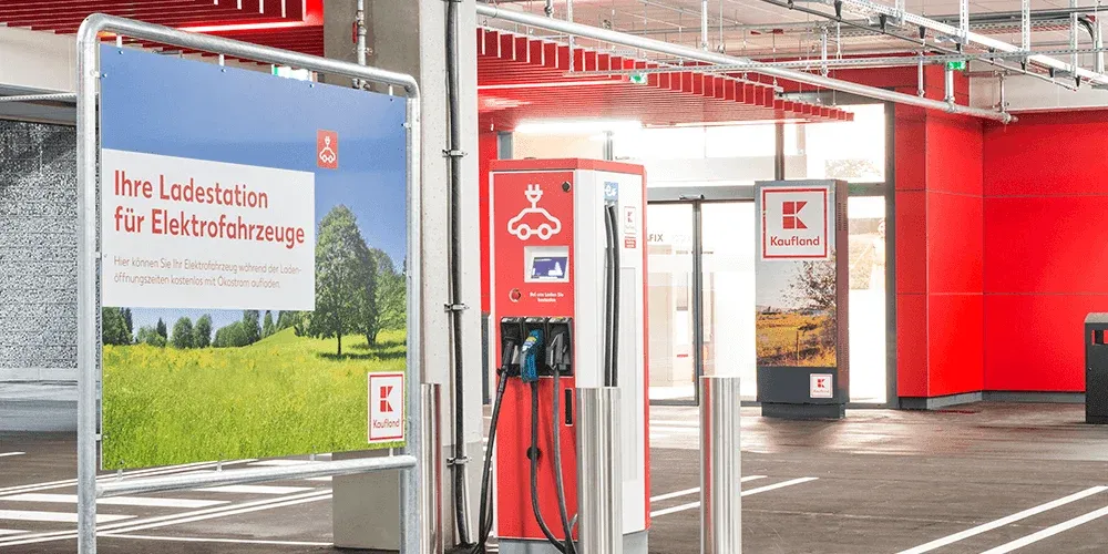 Electric vehicle charging station in a parking garage with signage in German, featuring red and white branding and a scenic photo.