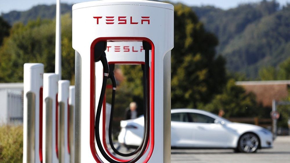 Stazione Tesla Supercharger con più unità di ricarica. Un'auto Tesla bianca è parcheggiata nelle vicinanze, circondata da alberi e colline.