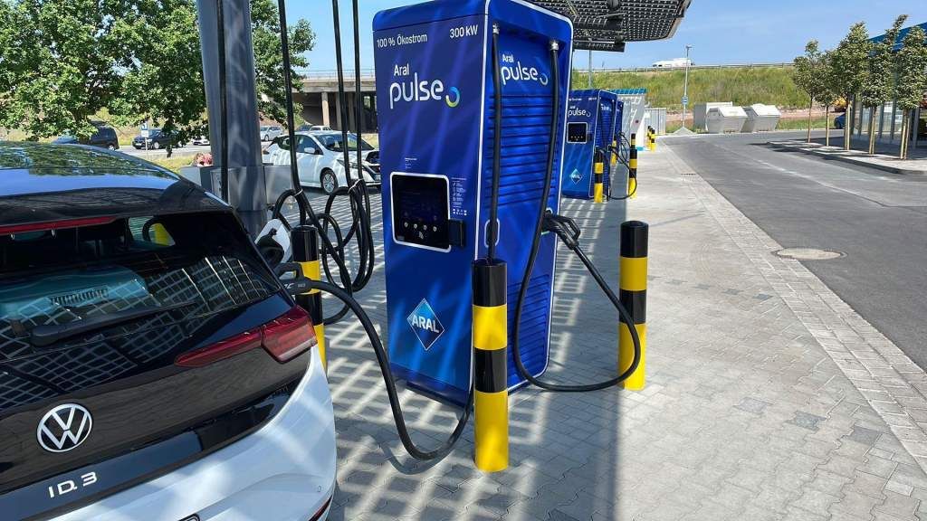 Aufladen eines Elektroautos an einer blauen Aral-Pulse-Tankstelle unter einem Sonnendach, mit mehreren Ladegeräten im Hintergrund an einem sonnigen Tag.