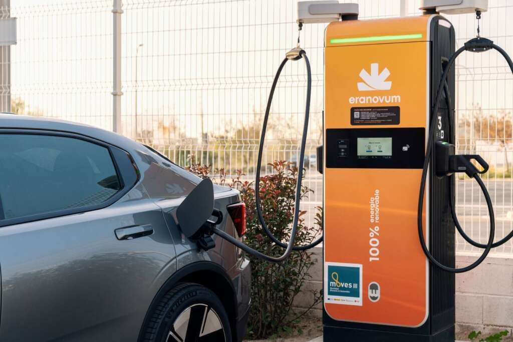 Carga de vehículos eléctricos en una estación de carga naranja denominada "eranovum" con vegetación al fondo.