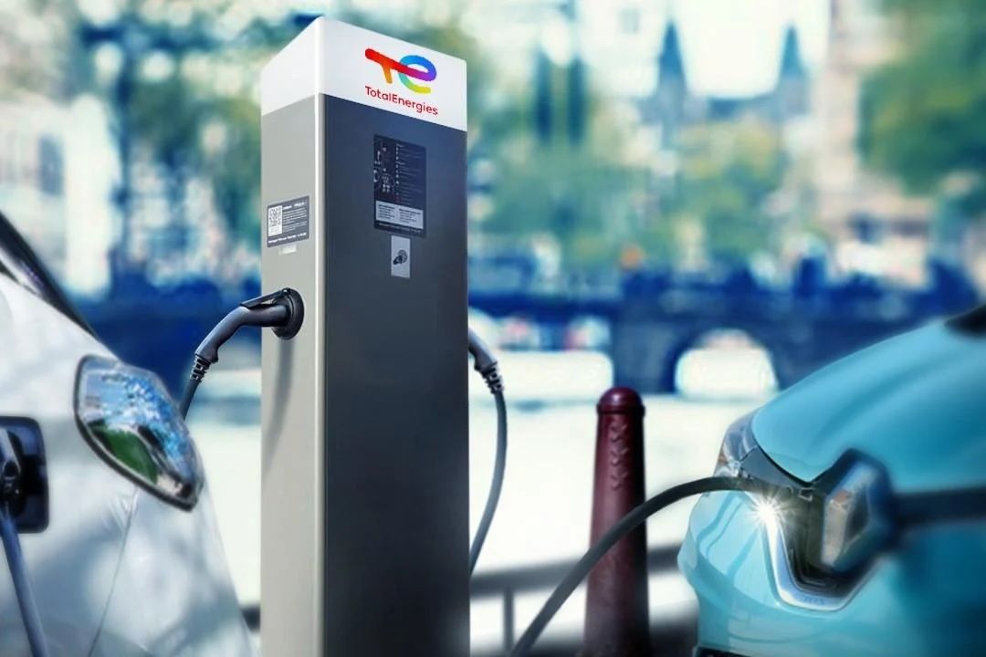 Deux voitures électriques en charge dans une station TotalEnergies. L'arrière-plan montre un paysage urbain flou avec des arbres et des bâtiments.