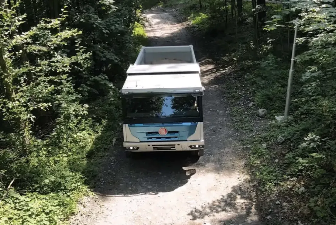 Un camion blanc et bleu roule sur un chemin de terre étroit et bordé d'arbres, dans une zone boisée.