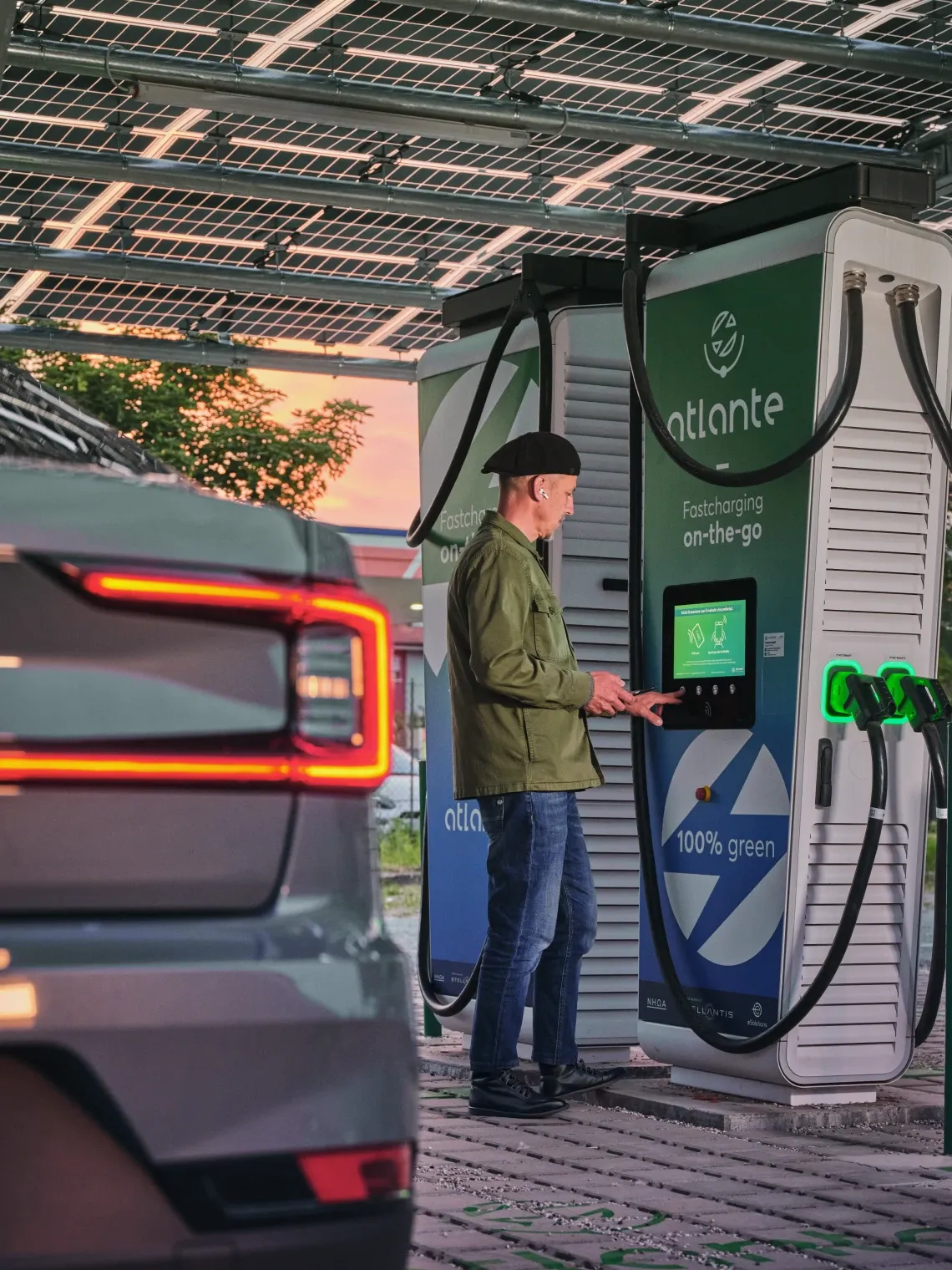 Al tramonto, una persona ricarica un veicolo elettrico presso una stazione di ricarica ad energia solare di Atlante, con un'auto in primo piano e il logo dell'energia verde ben visibili.