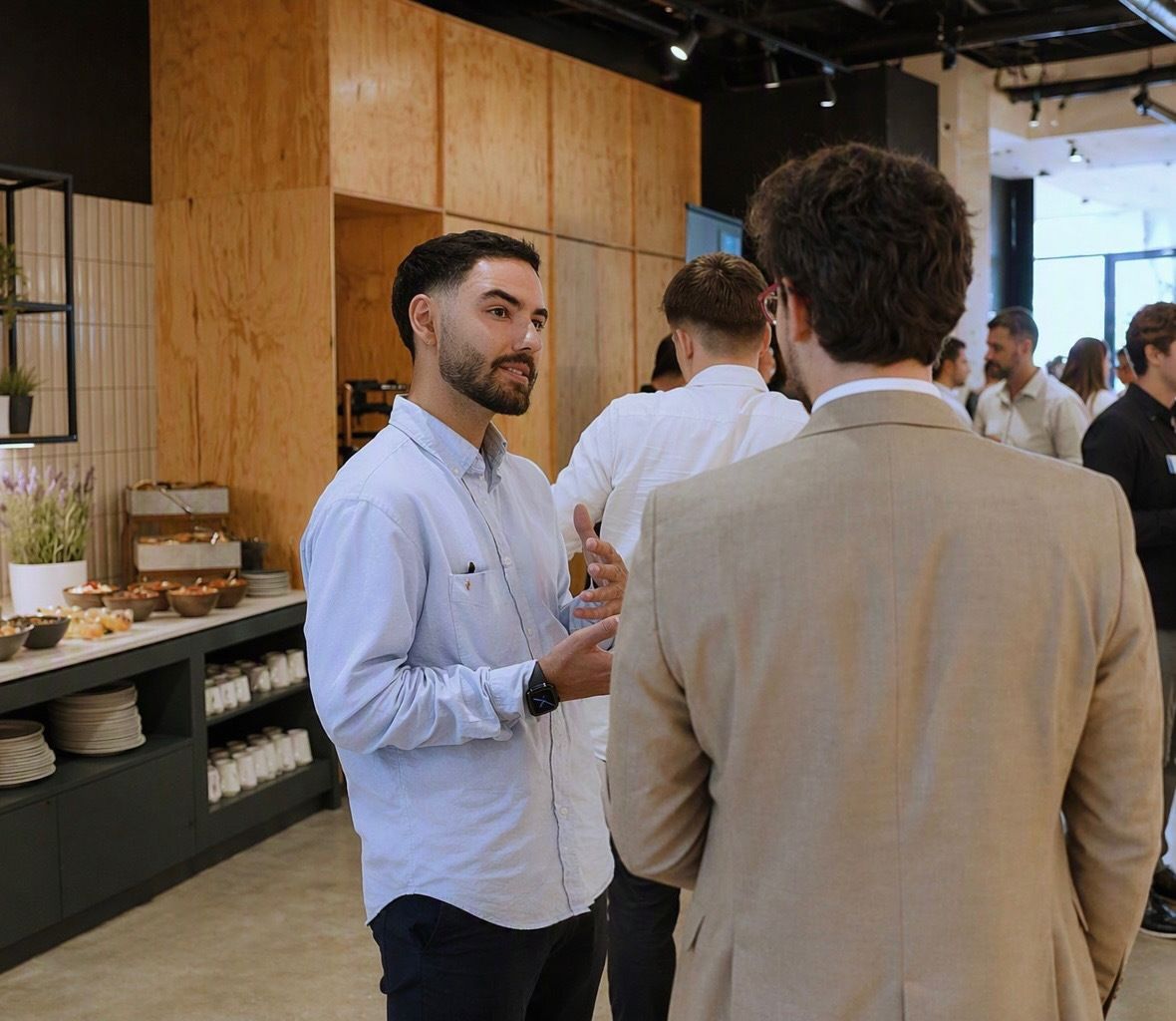 Two men in conversation at a networking event, with food and drinks on a counter in the background. Multiple people are mingling.