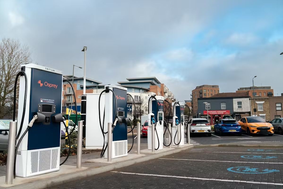 Stazione di ricarica per veicoli elettrici Osprey con più caricabatterie in un parcheggio, circondata da edifici e auto parcheggiate sotto un cielo nuvoloso.