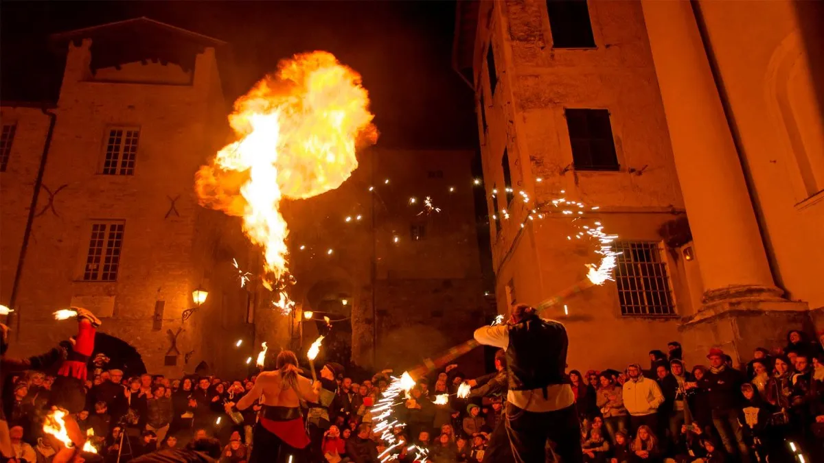 Uno spettacolo notturno con il fuoco in una piazza acciottolata, dove gli artisti creano palle di fuoco e scie di scintille, circondati da una folla incantata.