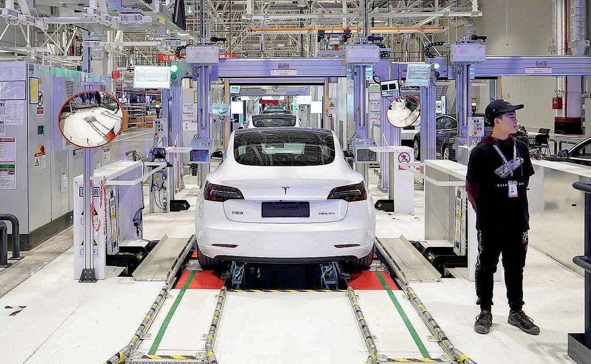 Un'auto elettrica bianca su una catena di montaggio in una fabbrica, con un operaio in piedi nelle vicinanze e vari macchinari e attrezzature intorno all'area.