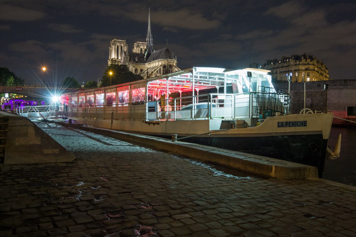 Illuminated boat docked on a cobblestone riverbank at night, with Notre-Dame Cathedral in the background.