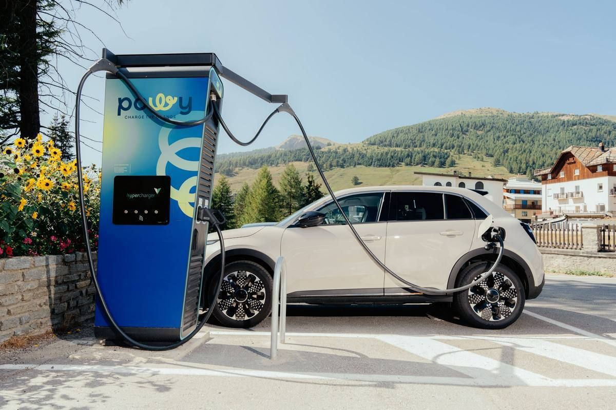 Ricarica di un’auto elettrica presso una stazione Powy, situata vicino a una strada con fiori selvatici, montagne ed edifici sullo sfondo, sotto un cielo azzurro e limpido.