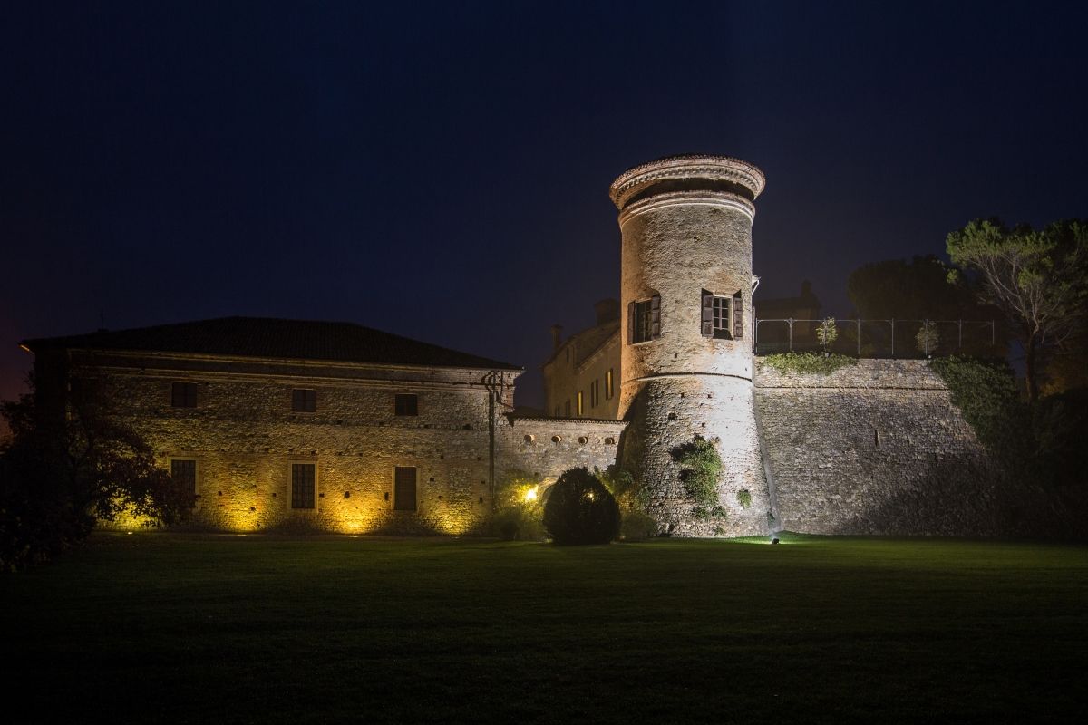 Castello Scipione. Uno storico castello in pietra con una torre rotonda, illuminato di notte, circondato da un cielo scuro e con un'area erbosa in primo piano.