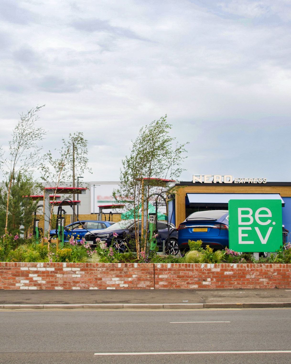 Electric vehicle charging station with cars parked, surrounded by greenery. A "Be.EV" sign and "Nero Express" building are visible.