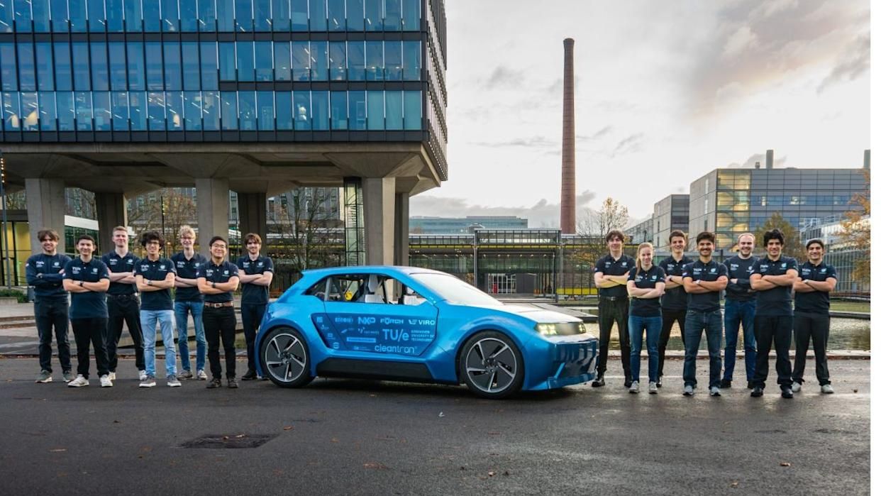 Team of people in matching shirts stand beside a blue electric car, with modern buildings and a tall chimney in the background.