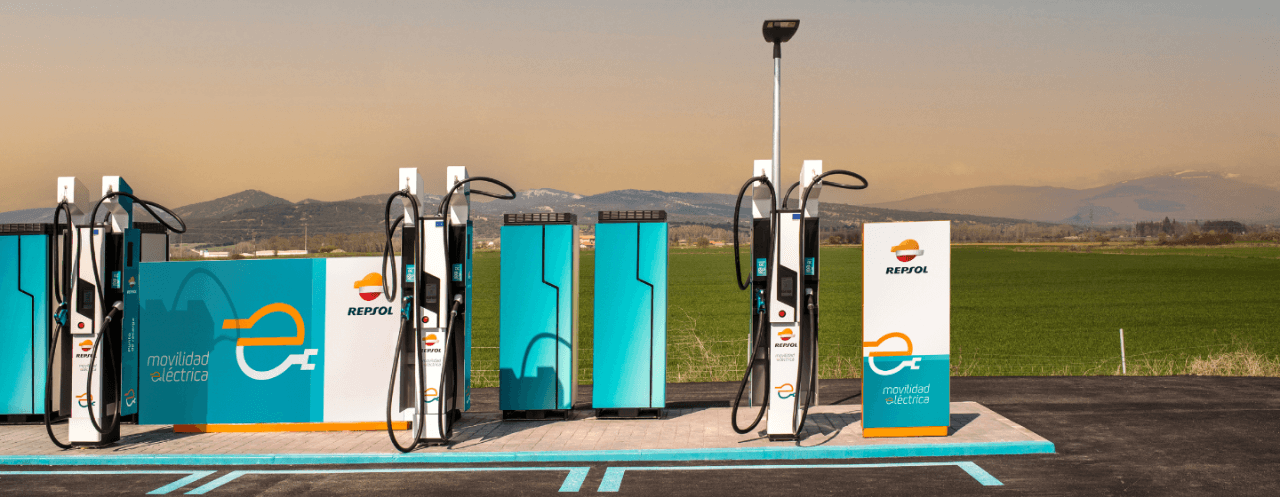 Estação de carregamento de veículos elétricos com vários carregadores, marca Repsol e um cenário pitoresco de campos e montanhas distantes.