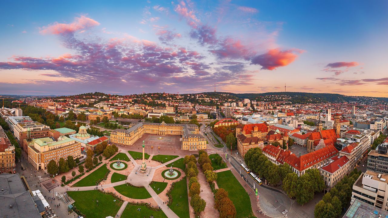 Luftaufnahme von Stuttgart bei Sonnenuntergang mit historischen Gebäuden, einem zentralen Park mit Springbrunnen und leuchtenden Wolken am Himmel.
