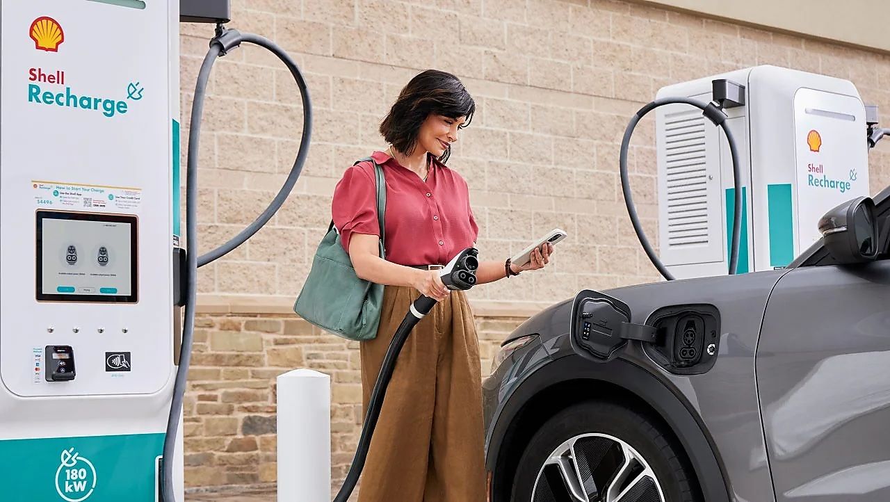 A woman charges her electric car at a Shell Recharge station, holding a charging cable and using a smartphone.