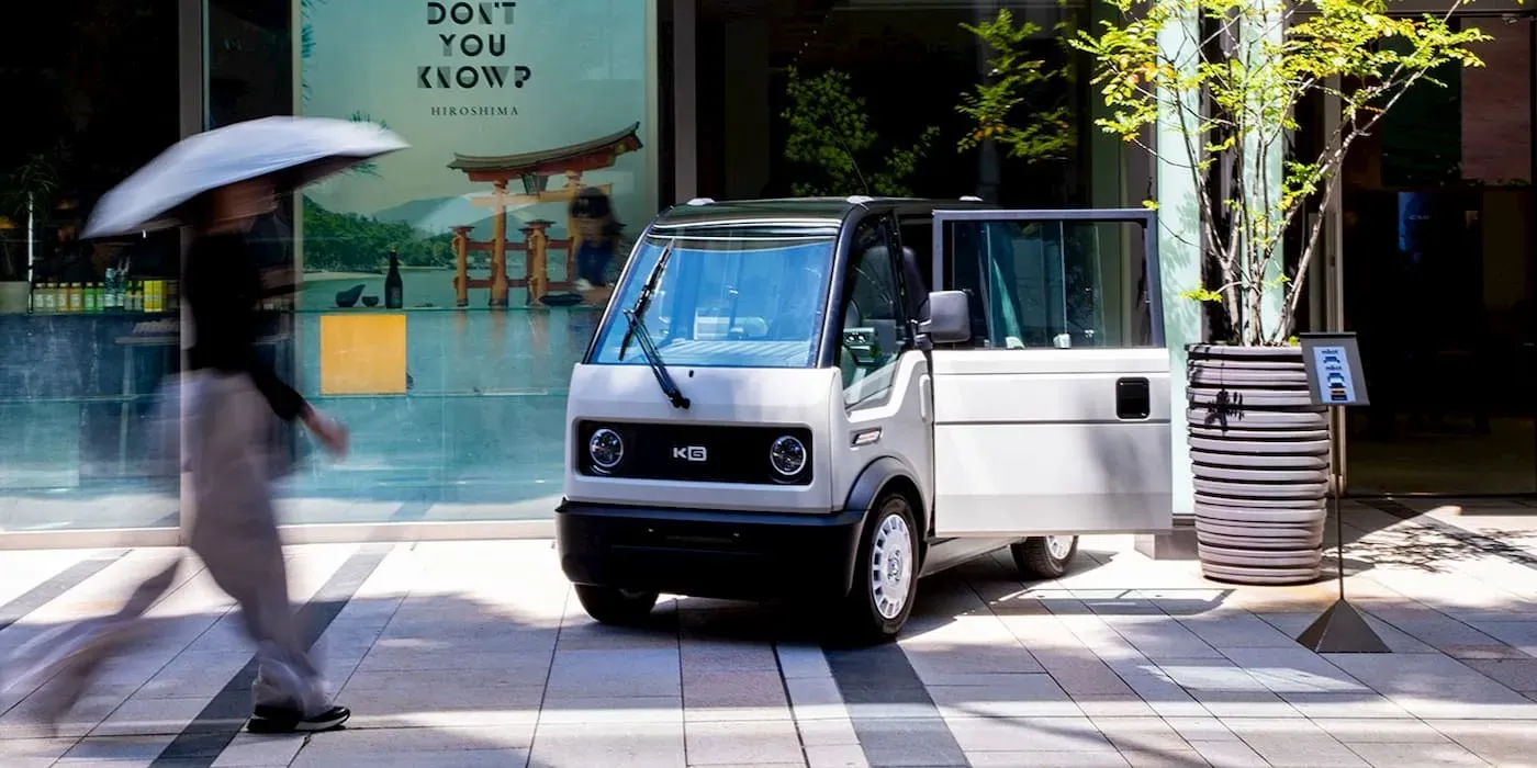 Un veicolo elettrico bianco compatto parcheggiato su una strada cittadina, con un pedone sfocato che cammina tenendo in mano un ombrello.