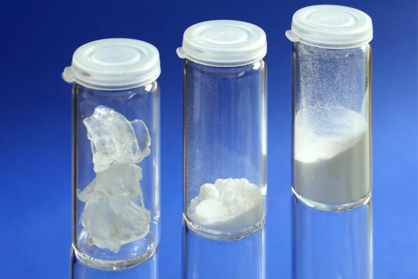 Drei Glasfläschchen mit weißen Substanzen: große Kristalle, kleine Körner und feines Pulver vor blauem Hintergrund.