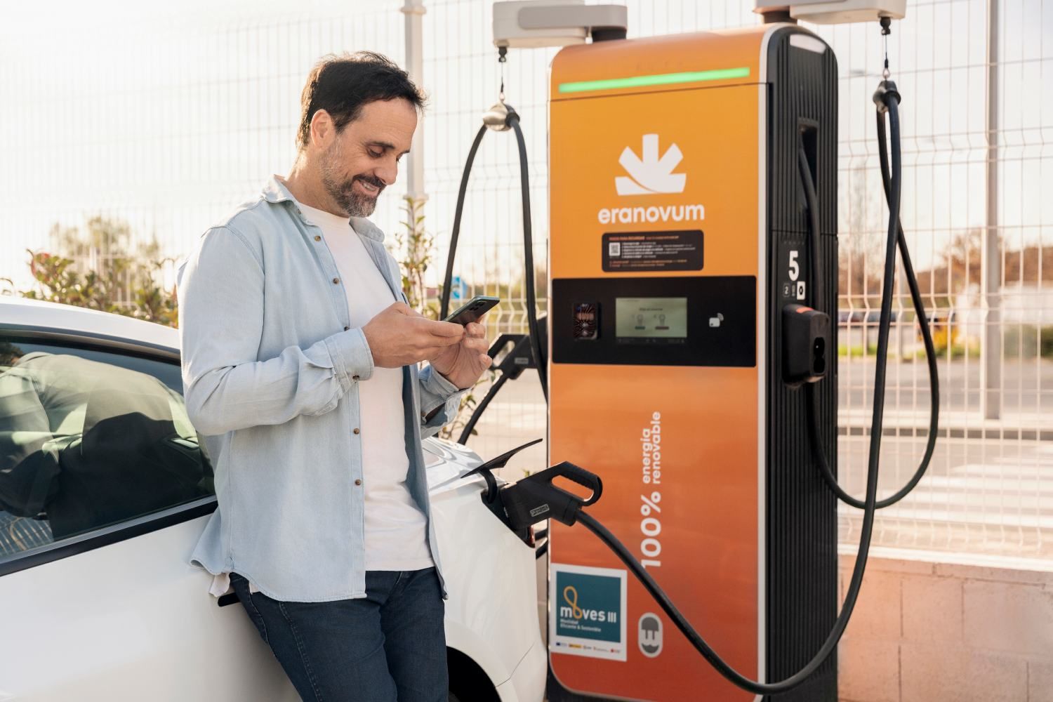 Hombre sonriendo al teléfono mientras carga un coche eléctrico en una estación de carga naranja al aire libre.