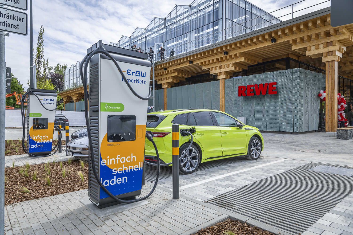 A lime green electric car charges at an EnBW HyperNetz station in front of a REWE store with modern architecture.