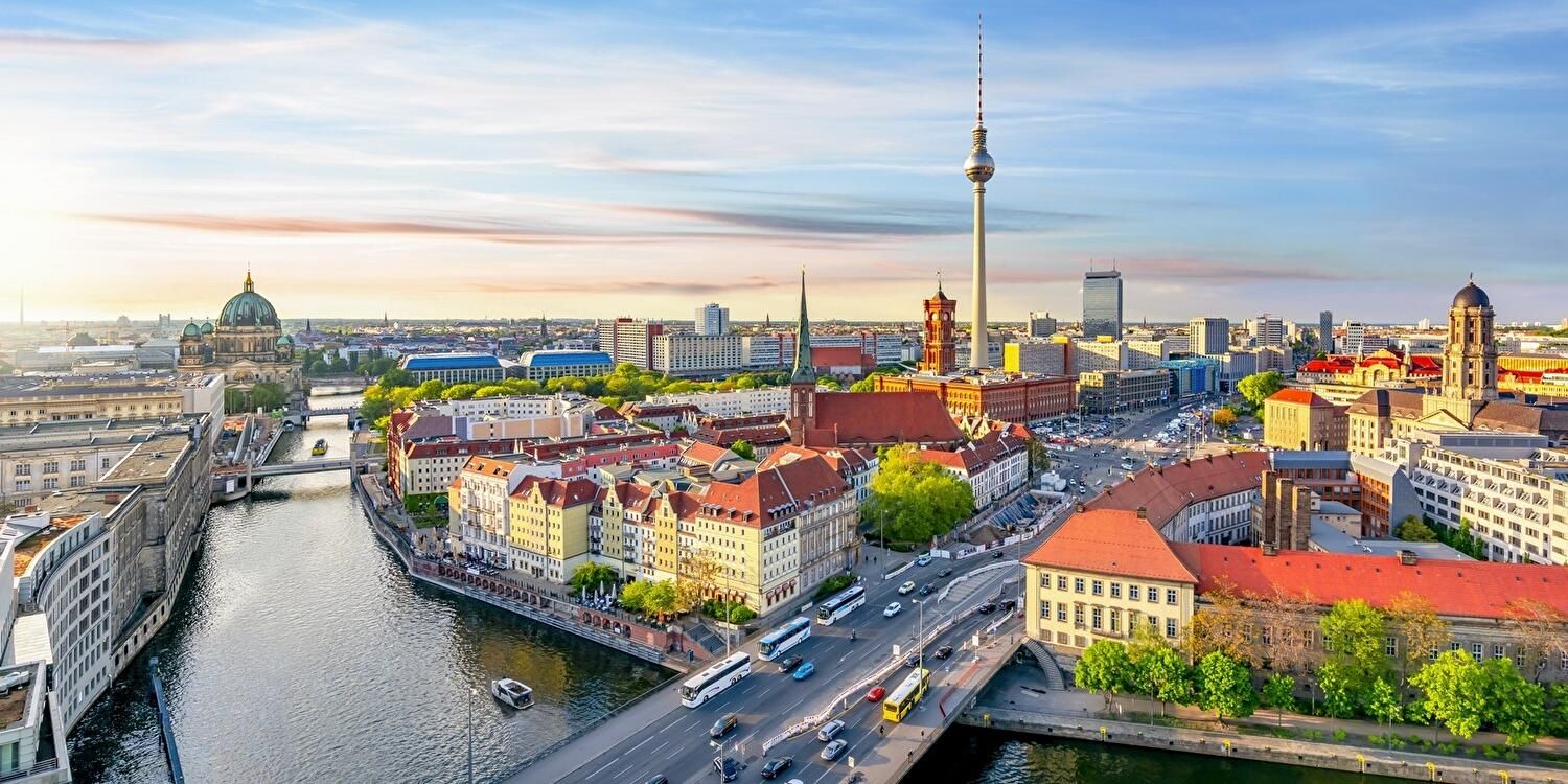 Eine Luftaufnahme von Berlin mit einigen historischen Gebäuden, einem futuristischen spitzen Turm und einem Fluss, über den eine Autobrücke führt.