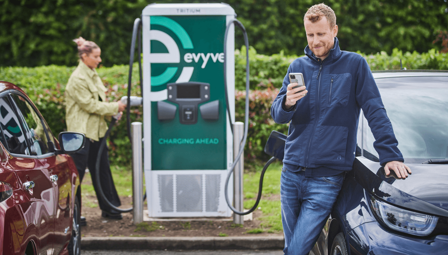 A man leans on an electric car using his phone while a woman charges a vehicle at a nearby station with "evyve" branding.