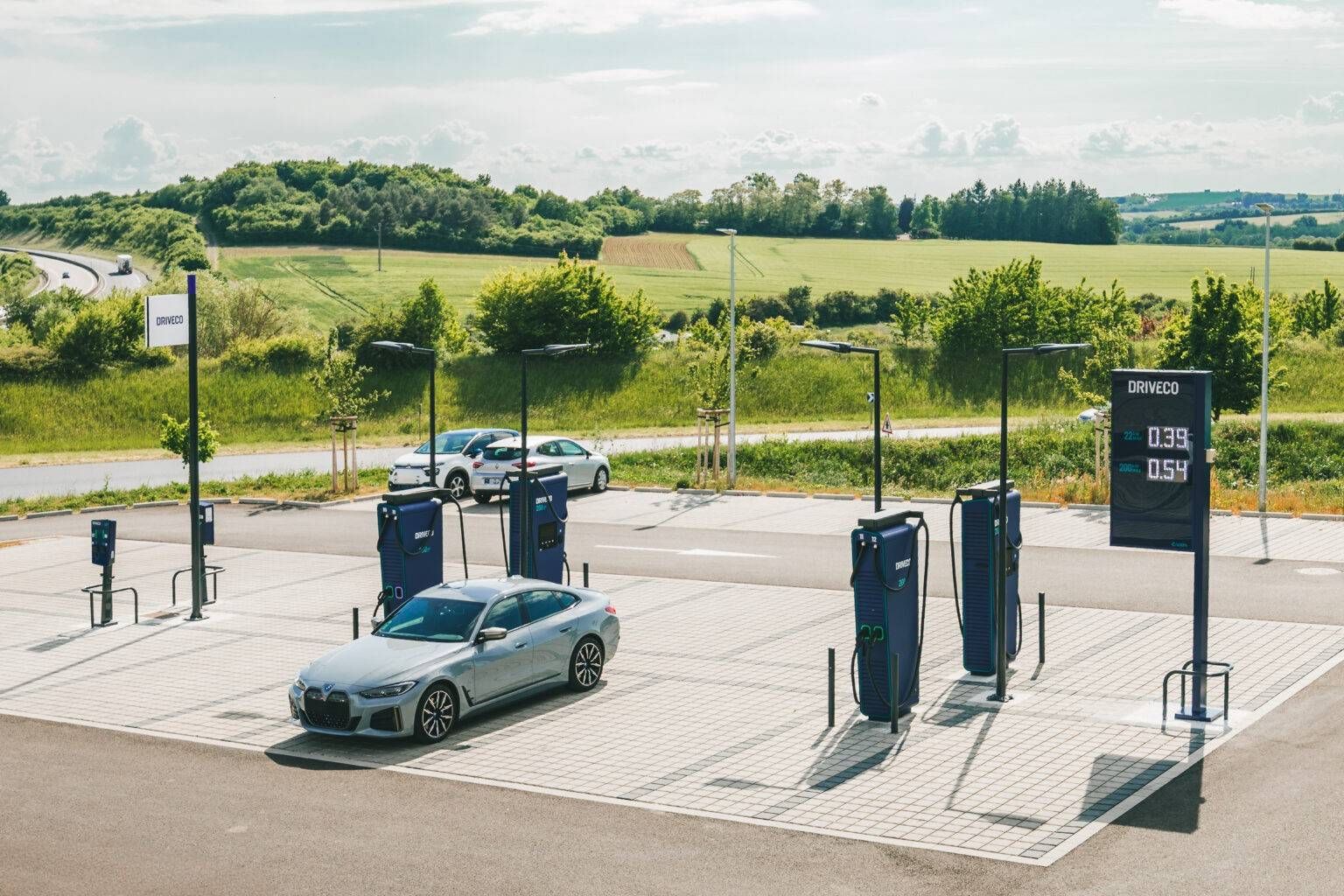 Une voiture argentée garée à une borne de recharge pour véhicules électriques dans une zone rurale avec des champs verts et un ciel bleu en arrière-plan.