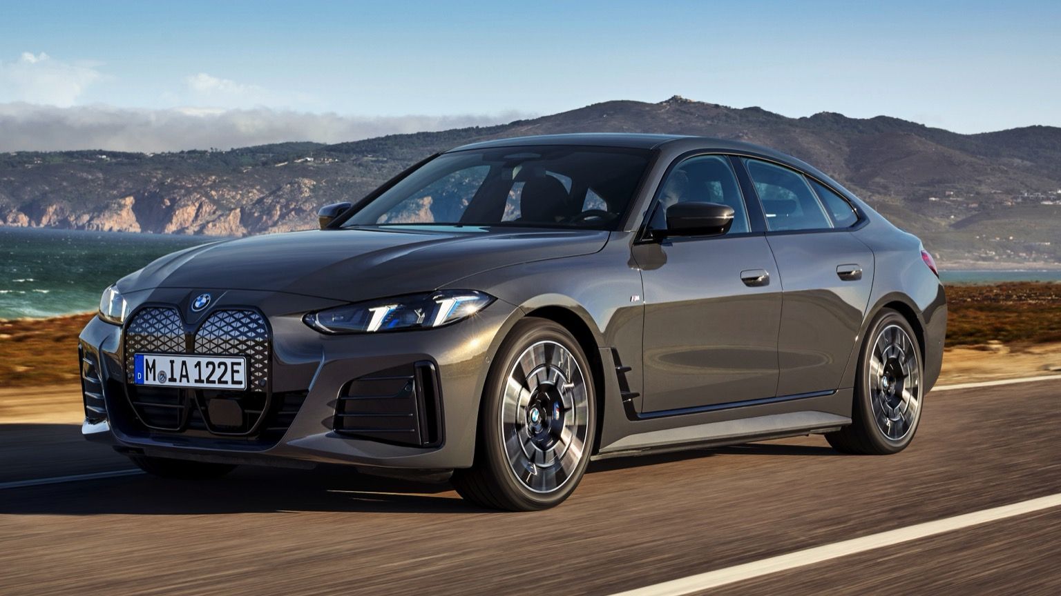 Un elegante BMW i4 gris circulando por una carretera costera con montañas y océano de fondo bajo un cielo azul claro.