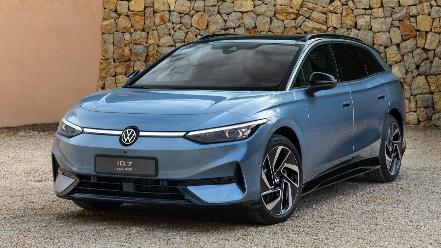 Volkswagen ID.7 Tourer blu parcheggiata su ghiaia, con uno sfondo di muro in pietra. Caratterizzata da un design elegante e fari moderni.