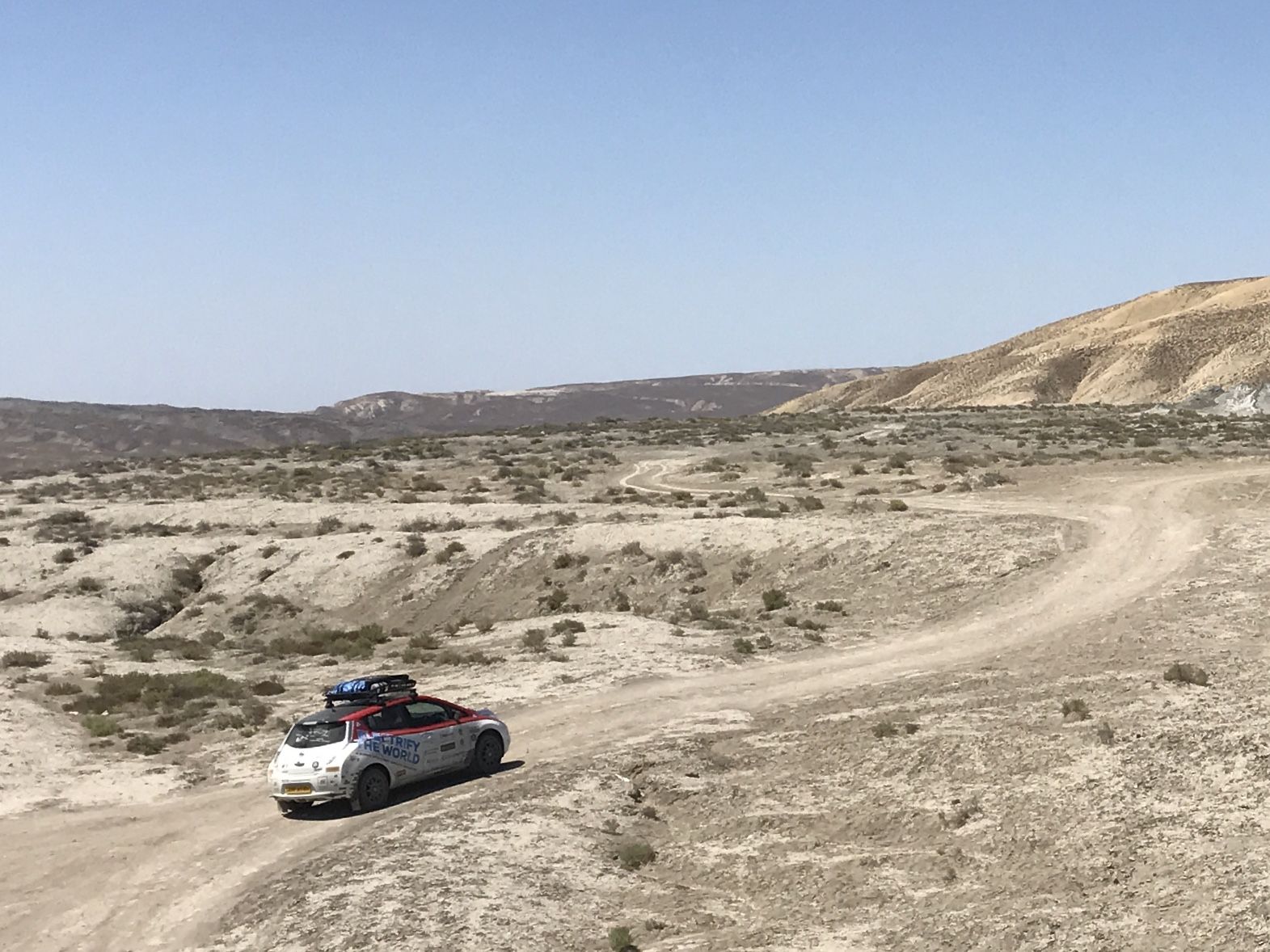 Un'auto con bagagli sul tetto percorre una strada sterrata tortuosa attraverso l'Azerbaijan sotto un cielo azzurro e terso.