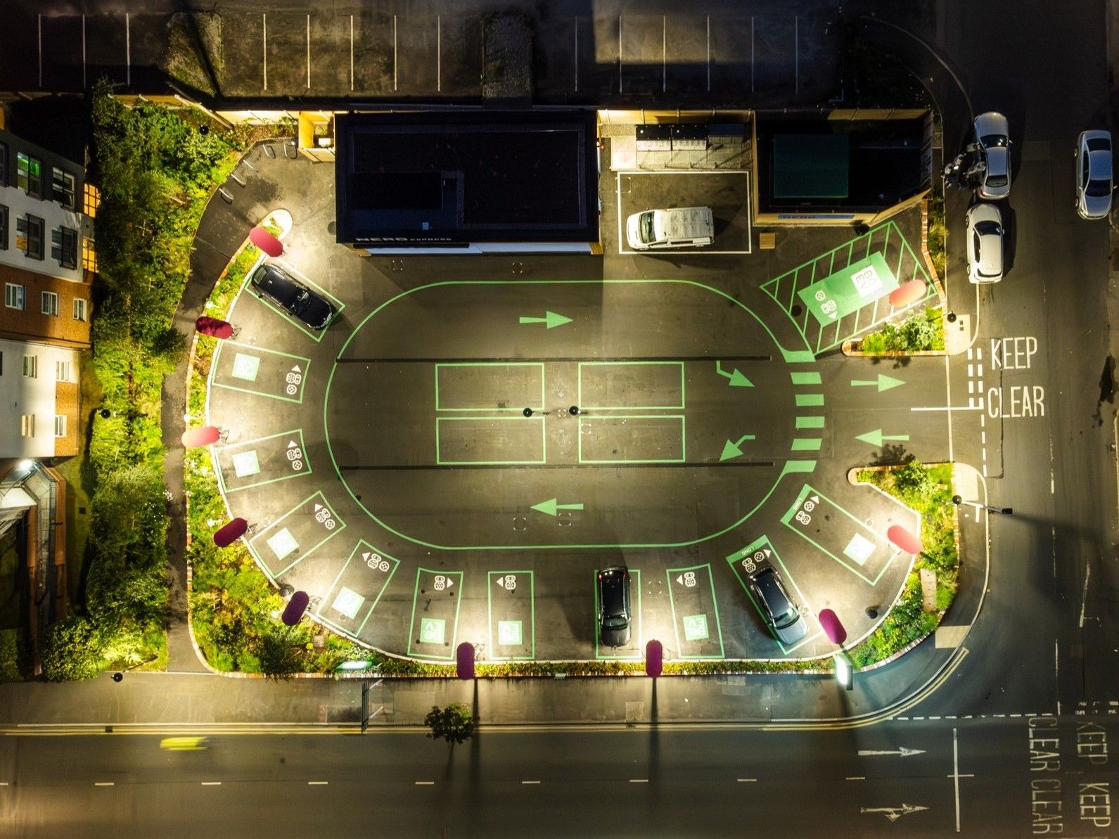 Aerial view of a well-lit, oval-shaped urban parking lot at night, with cars parked in marked spaces with electric vehicle chargers and surrounding greenery.