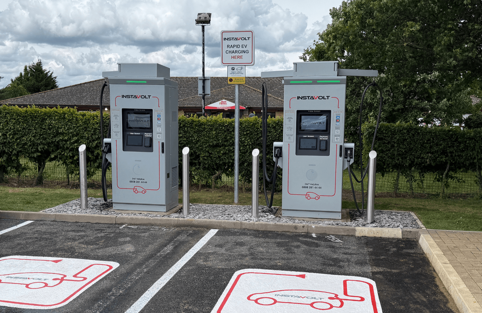 Two grey Instavolt fast chargers at the back of two parking spaces with symbols on the ground showing a car charging with Instavolt's logo inside. The chargers are in front of green hedges with a tiled roof and grey sky in the background.